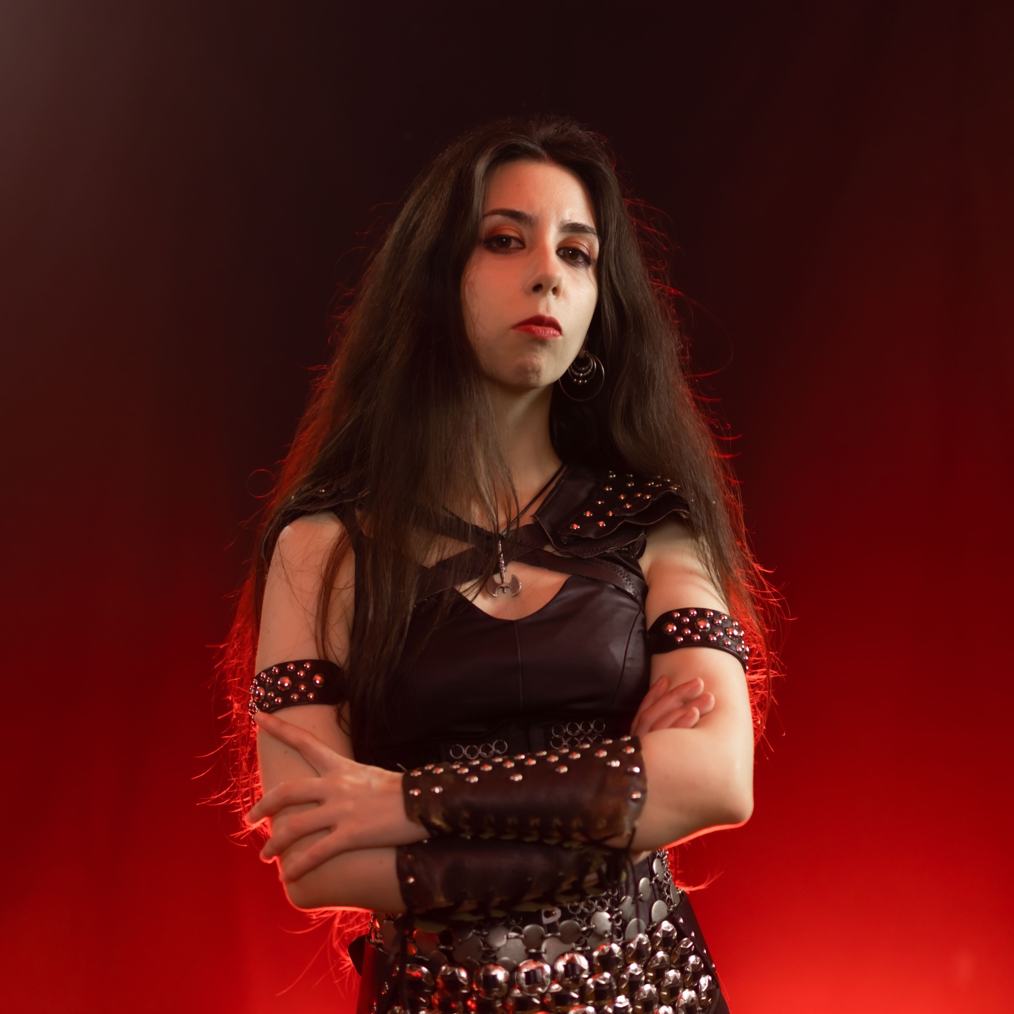 Mina Walkure, Spanish vocalist of the heavy metal band Bronze, posing with arms crossed in studded leather armor against a red-lit studio background. Photo by Emil Westin, edited for the Realm of the Damned photoshoot session.