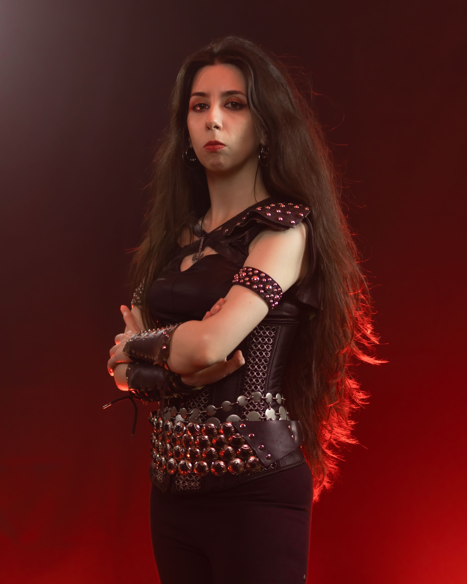 Mina Walkure, Spanish vocalist of the heavy metal band Bronze, posing with arms crossed in studded leather armor against a red-lit studio background. Photo by Emil Westin, edited for the Realm of the Damned photoshoot session.